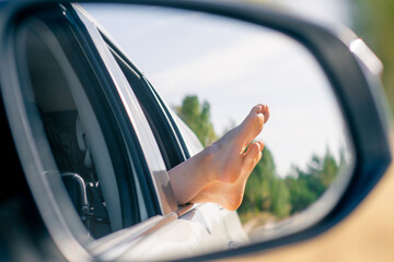 In the side mirror of a car the reflection of a woman’s legs stretched out of window onto the street while driving through field - Powered by Adobe