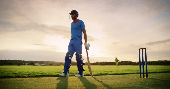 Portrait of a Professional Cricket Player in Blue Uniform and Protective Helmet Waiting for the Game to Start, Standing with a Bat on a Field. Cinematic Low Angle, Warm Sunset Scenery Footage