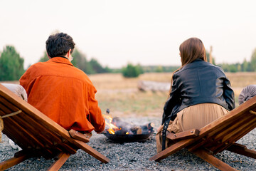 Back shot of young man and woman sitting in nature by fire relaxing and roasting marshmallows