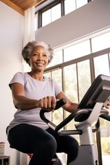 Fototapeta premium Vertical photo of a senior black woman during workout on a smart exercise bike at home. She smiling and looking at camera. A scientific approach to training for maximum performance.