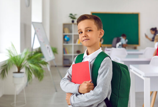 Proud boy first grader with satchel and workbook posing on first day of elementary school getting ready to receive quality assignment standing in classroom with big blackboard. Back to school