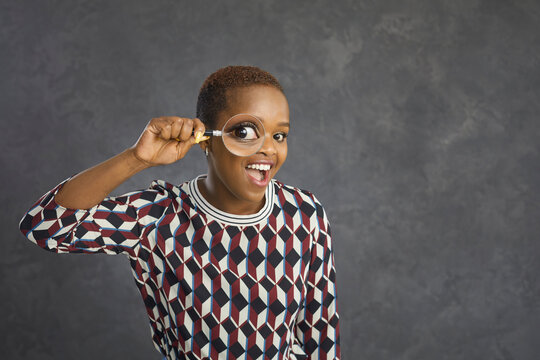 Studio shot of happy black woman with magnifying glass looking at you with funny big eye. Young lady looking for something interesting, searching for fascinating wow facts, making amazing discoveries