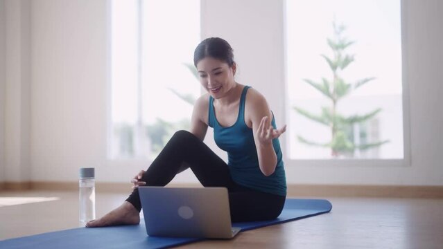 Asian female fitness trainer in workout is conducting online fitness classes through video call on her laptop to help students maintain their strength and good health while exercising at home.