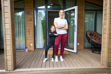 Mother and pretty little daughter standing against cozy country house