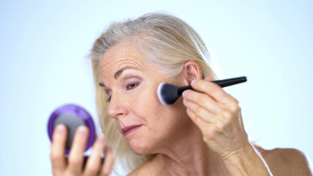 Closeup of blond gray hair woman looking in compact mirror, putting powder on her face with brush, then looking at camera. Studio white background.