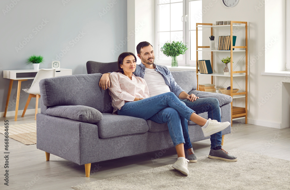 Wall mural happy relaxed young family couple sitting on sofa at home. smiling husband and wife watching televis