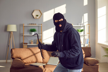 Burglar with crowbar in the house. Robber man wearing black jacket, balaclava mask and gloves breaks into apartment. Sneaky thief walking on tiptoes and looking around for valuable stuff he can take - obrazy, fototapety, plakaty