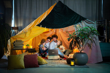 In front of the camera happy parents together with their kids watching movie together on the laptop while sitting down on the floor inside the extempore tent on the living room and eating popcorn - Powered by Adobe