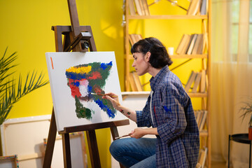 A close up shot of a woman dipping her brush into her work art palette and painting onto a portrait while being very concentrated