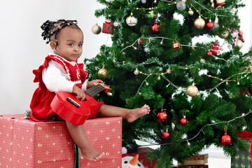 Adorable happy smiling African little girl child sitting on big gift box present under Christmas tree in living room, cute kid celebrating winter holiday. Merry Christmas and Happy New year concept.