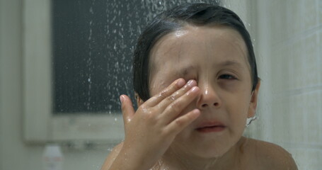 Child touching eye with finger while underneath of shower head with water flowing , rubbing face...