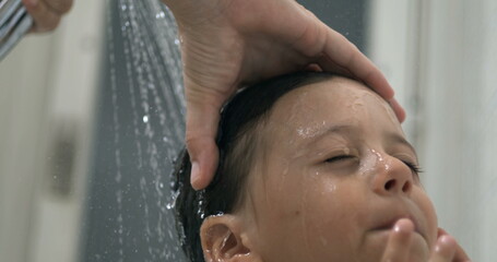 Close-up child face underneath flowing water shower head during bath time routine, young boy with...