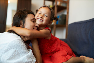 Side view of cute happy smiling little girl in red dress hugging closely her mother on sofa, demonstrating affection, woman cuddling with kid, showing unconditional love for her child - obrazy, fototapety, plakaty