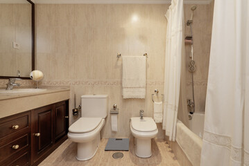 Bathroom with cream-colored marble-like tiling, bathtub with curtains, mahogany-colored wooden vanity unit and mirror with matching frame on the wall