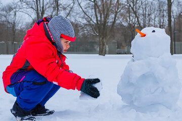 Fototapeta na wymiar boy makes a snowman. a little boy walks outside in winter and makes a big snowman out of white sticky snow, a boy walks down the street in winter