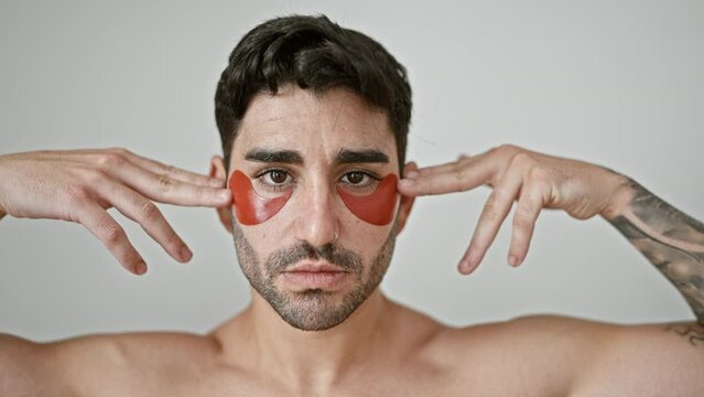 Young hispanic man standing shirtless putting baggy eyes patches over isolated white background
