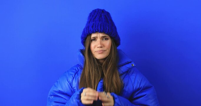 Angry sad brunette young woman in blue puffer and knit hat posing isolated on blue background studio. Woman shout screaming news with hand near mouth shocked wow.