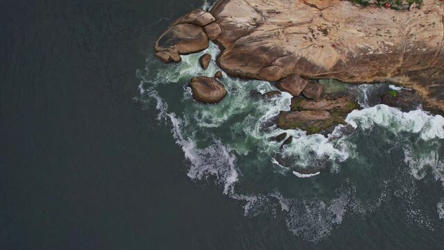Drone Footage of Ipanema Beach in Rio De Janeiro in Brazil. The video is a 90 degree down view of the pedra do arpoador with waves crashing on it.