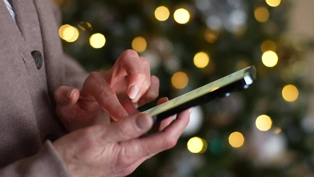 Closeup side view of unrecognizable joyful woman in room tapping and texting on smartphone wishing congratulation on xmas Eve, on blurred background bright bokeh lights of Christmas tree, slow motion.