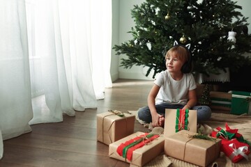 A boy with long blond hair in a white T-shirt and red plaid pants under a Christmas tree in a room...