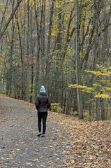 Fototapeta premium woman walking on a hiking trail in Taughannock Falls State Park (waterfall, cayuga lake near ithaca, upstate new york) in autumn with fall foliage (leaves changing colors) wearing brown jacket, hat