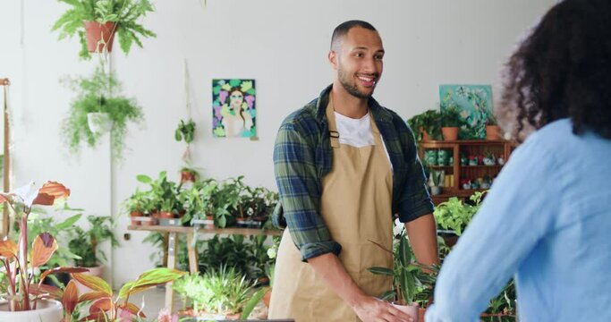 Cheerful handsome young man florist selling flower pot. Woman buys spring housplants in flower store. People in flower shop, client pushing cart. Buyer talking to sales gardener in greenhouse.