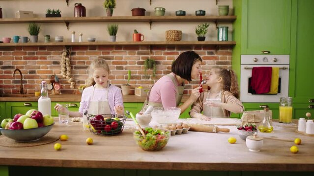 Singing with a tablespoon happy mother while preparing some delicious biscuits with her two charismatic daughters together very happy they smiling large at the kitchen island