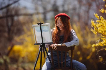 Tuinposter Beautiful girl with red hair painting on a canvas while smiling and sitting at the park © qunica.com