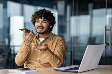 Cheerful and smiling successful businessman using app on phone to record audio message, Indian man...