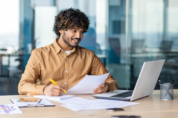 Young successful financier behind paper work inside office, satisfied smiling man works with laptop...