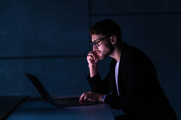 Young business man working on computer at night in dark office. The designer works in the later...