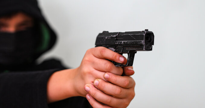 A hooded young man in black aims a gun. Concept of crime and gun attack with young man. Armed robbery on black, shallow depth of field, focus on gun barrel.