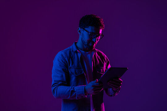 Young man with modern digital device isolated over gradient blue studio background in pink neon light.