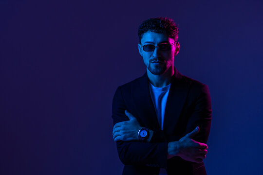Young man with beard wearing sun glasses and a leather jacket between red and blue neon lights