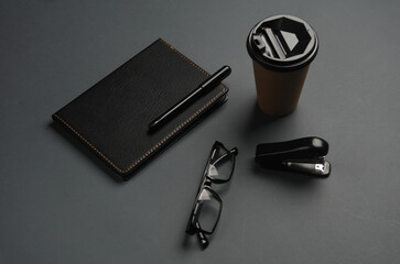 Notepad, stapler, eyeglasses, coffee cup on dark gray background. Black stationery objects