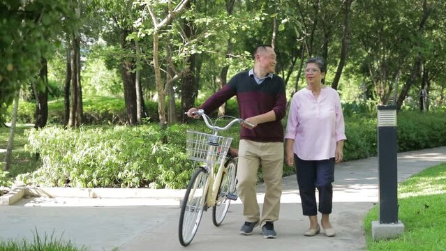 An elderly Asian couple walks with a bicycle in a park. Living happily in retirement. Elderly health care concept