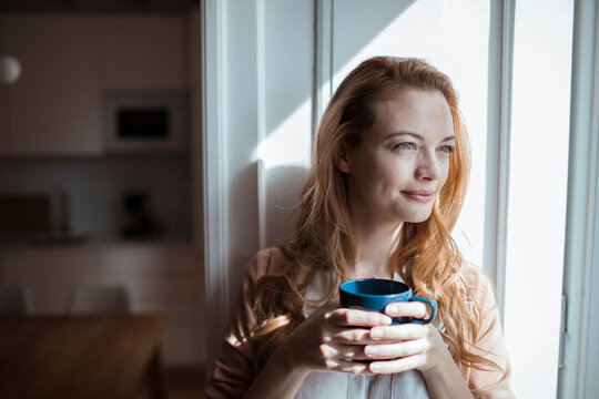 Young woman looking out the window while having coffee at home