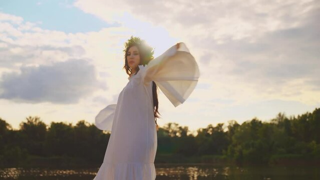 happy young beauty woman smiling face dancing by river bank water sun light blue sky green forest. Fantasy girl raised hand to sky nature. herbal wreath long hair white dress fly in wind slow motion