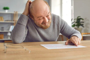 Senior man with dementia gets confused and scratches head when he looks at calendar on table. Old...