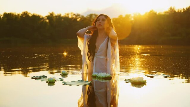 fantasy woman river nymph stands in lake, hand gently stroking touching long healthy dark hair water lily flowers. Sexy girl enjoy summer nature sun light magic sunset tree forest. natural cosmetics