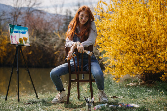 Beautiful ginger girl smiling while sitting and holding art supplies in her hands at the park