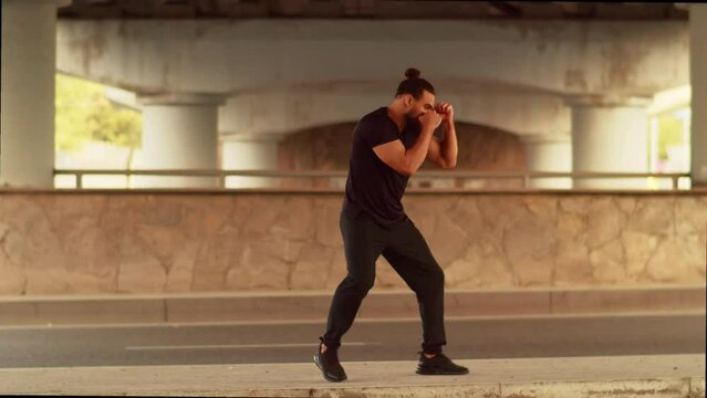 Young muscular sportsman boxing with a shadow and working out dodges on a sidewalk under the bridge in the city. Concept of modern outdoor sports training in a casual urban life