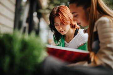 Two high school girls study together outdoors, casually dressed, reading and discussing school...