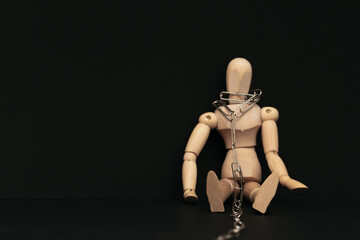 Wooden man sitting on a dark background with a chain around his neck. The concept of non-freedom.