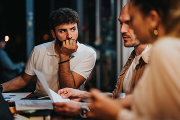 Confident business partners discuss project details over documentation at a coffee bar. The young executives analyze profitability, plan strategies, and work together remotely.