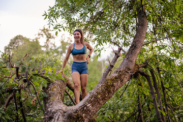 Naklejka na ściany i meble An active and attractive girl warms up in a park, flexing and exercising on a tree. She enjoys a fit and healthy outdoor lifestyle, engaging in sports amidst nature.