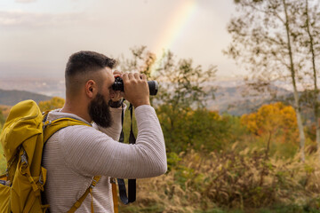 With a backpack on and wearing a sweater, a hiker gazes through binoculars, a rainbow visible in the distance following the rain.