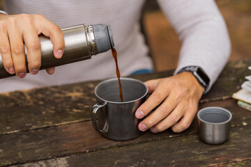 Close-up of hands as a man seated at a mountain table pours coffee, a well-deserved break after a hike..