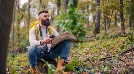 During his woodland hike, a hipster enjoys a rest, sitting on a tree stump while consulting a paper map to plan his route.