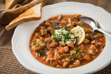 Ost Deutsche DDR GDR Soljanka Eintopf Suppe mit Jagdwurst, Saure Gurken, Letscho, Schmand, Zitrone, Toast und Petersilie mit L&ouml;ffel, Salzstreuer und braun Stoff Hintergrund